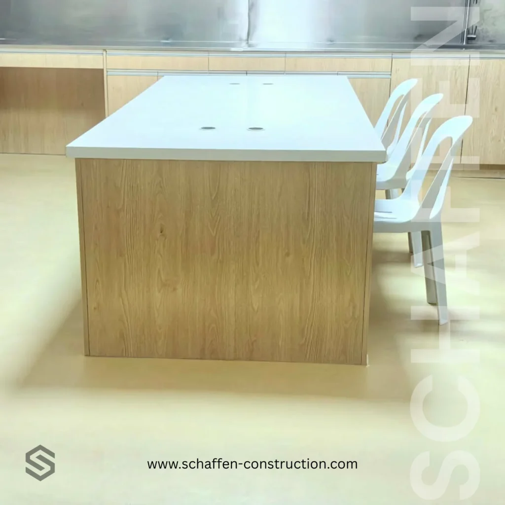 Modern kitchen showroom display: wooden island with a white countertop and built-in outlets, accompanied by white chairs along the side.